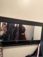 Black wall mirror mounted on wall with four hooks visible, holding coats. Mirror surface reflects person taking photo and coats hung behind.