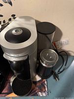 Silver and black Nespresso coffee maker with attached water reservoir and drip tray, alongside a black electric milk frother with silver top and clear lid. Both have power cords visible.