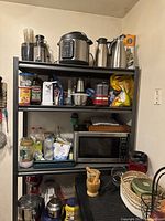 Photo of metal open shelving unit with four shelves filled with kitchen appliances, bottles, jars, and other items, against a beige wall.