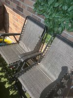Two beige-grey fabric sling outdoor lounge chairs with black metal frames, armrests, and adjustable reclining backs, placed against a brick wall and greenery.