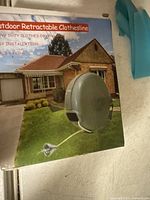 Outdoor retractable clothesline in grey plastic housing with metal line, shown in box with product packaging featuring house image.