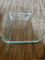Clear square Pyrex baking dish with integrated handles, placed on a carpeted surface.