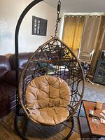 Front view of the brown wicker hanging egg chair with tan cushioned seat and black metal stand in living room.
