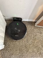 Front and top view of black Ultenic robotic vacuum cleaner on a carpeted floor with charging dock plugged into the wall, showing illuminated green power and charging icons on the vacuum.