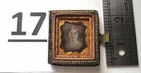 Full view of the closed daguerreotype case with black and gold floral design, showing the female subject visible through the glass on the front mat.