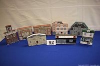 Front view of all eight wooden miniature buildings displayed in a row on blue surface, showing detailed facades.