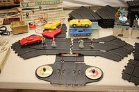 Full shot of assembled portion of slot car track with red Ferrari 330 P/LM, yellow Testarossa and blue Chevy Impala cars on display, flagman figures, and controllers visible