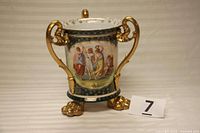 Full view of the three-handled porcelain loving cup showing gilded handles and lion's paw feet with decorative scenes on the body.