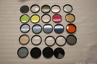 Top-down view of 20+ assorted colored 49mm camera lens filters arranged in rows on neutral surface.