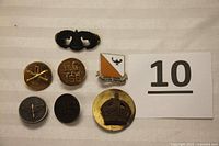 Seven military lapel pins displayed on a white surface, showing varied designs including crest, wings, crown, and 'US' insignia.