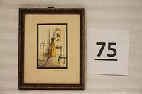 Framed hand-colored photograph showing woman in yellow dress looking into a mirror on a console table, with signature visible on mat.