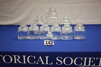 Ten clear glass storage/display jars with ground glass lids arranged in three rows on a blue surface starting with the smaller jars at front and largest at back; jars are square shape, two larger ones taller and rectangular.