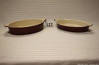 Two oval burgundy and white ceramic casserole dishes placed side by side on a white cloth backdrop with a lot number label (122)