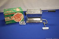Full view of chrome pasta machine, detachable crank visible separated from machine, on blue cloth background.