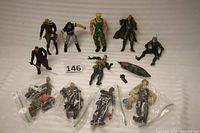 Top view of nine Metal Gear figures and one WWF wrestling figure arranged on a white surface with some accessories and weapons in plastic bags below them.