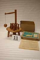 View of the wooden tabletop skittles game showing the round table with three legs, nine pins, swinging ball on string, original box, and instruction sheet.
