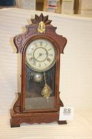 Full front view of antique New Haven clock showing wooden case with glass door, brass female head ornament on top, Roman numeral clock face, pendulum, and bell inside.