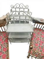 Front view of white metal miniature rack with three shelves and decorative curly metalwork, placed on pink floral cushion background.