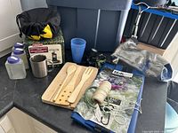 Full view of assorted camping items including Coleman stove box, plastic containers, mug, wooden utensils, dry bags, rope, and bungee cord on a countertop.