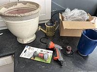 Wide view of the ceramic pot, decorative solar watering can, garden hose spray gun, packaged hose connector, and other hose parts on a countertop.