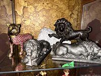 Overview photo of four animal figurines on shelf: black cast-iron lion, silver-toned lion, white ceramic lion head, and pink-spotted wild cat figurine.
