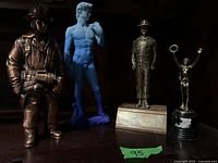 Four statues on a shelf: copper firefighter, blue David, soldier/park ranger, small trophy