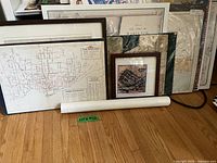Photo showing multiple framed and unframed maps and posters leaning against a wall, with visible rolled map in front.