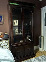 Full view of dark brown mahogany glass display cabinet with two glass doors and visible items inside, lower cabinet doors below, situated near a bed and other furniture.