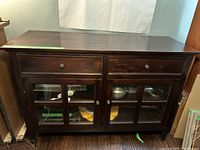 Overall front view of the dark brown wooden sideboard with two drawers above glass paneled doors, showing metal knobs and some contents inside.