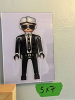 Glossy photograph of Playmobil style figure in black and white outfit with silver hair and sunglasses, 5 x 7 inches