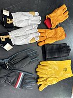 Photo showing all five pairs of gloves together: orange leather gloves with red knit cuff, white leather ski gloves with brown detail, black wool and leather Paul Smith gloves, black leather gloves with red, white and blue stripes on cuff, and yellow rubber heavy duty gloves.
