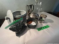 Various kitchenware items on white surface including new Le Creuset skillet with original label, glass and silver plated pitchers, stone bowl with lid, and bag of decorative rocks.