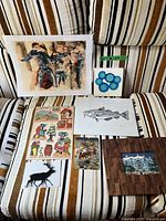 Full view of assorted art and postcards lying on striped fabric, showing several prints and posters including cod fish print, abstract circles, and others.