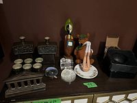 Overview of all items in the lot on dark wooden surface under lighting, showing the diversity of decorative objects including figurines, cups, bottles, glass pieces, and a black box.