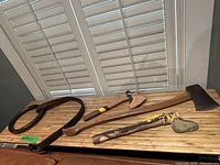 Lot contents on wooden countertop under window with plantation shutters: three axes/hatchets with wooden handles and leather sheath on one, alongside large iron tool