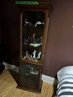 Full view of wooden curio cabinet with glass front and sides, wire mesh panel on lower door, standing on hardwood floor near bed.