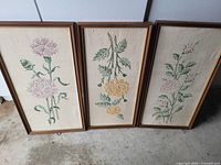 Three framed needlepoint floral pictures showing detailed embroidery on beige fabric with wooden frames in brown finish.