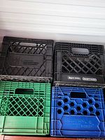 Four plastic milk crates stacked in two rows. Top: Two black crates with dairy branding. Bottom: One green and one blue crate with dairy branding and patterns.