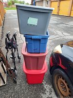 Four stacked plastic storage totes with lids in gray, blue, brown, and red colors; photo taken outdoors showing condition and colors.