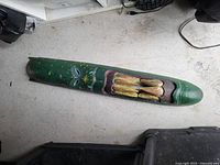 Long abstract face carved wooden mask with green base and yellow and purple painted accents, visible paint wear and minor scratches.