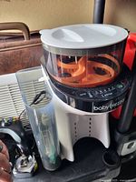 Baby Brezza infant formula maker showing the main unit with clear container and orange spiral mixing component, white base with black control panel and water tank attached.