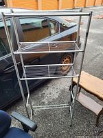 Photo of a metal standing bathroom shelf with three wire shelves set outdoors next to a black car and a wooden bench.