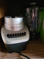 Photo of a Black & Decker blender on a wooden shelf. Shows base with control buttons labeled 0-6 and the clear plastic jar with lid and handle next to it.