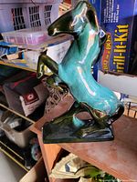 Glossy ceramic horse figurine in a rearing pose, turquoise, green, and black color tones, placed on wooden surface with boxed items in background