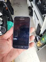 Photo showing front view of a Samsung smartphone with black screen held in hand. Device has physical home and navigation buttons.