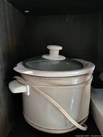 White electric cooking pot with glass lid and electric cord wrapped around it inside a cabinet.