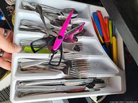 Overhead photo showing a cutlery tray containing forks, spoons, knives, colorful plastic handled utensils, kitchen scissors, and metal tongs.