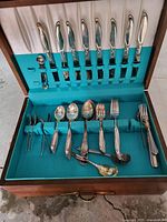 Silver tone cutlery set with knives, forks, spoons and serving pieces neatly arranged in a teal-lined wooden storage box.