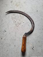 Photo of a hand sickle showing a curved metal blade with rust and a wooden handle on a concrete floor.