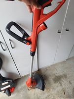 Full view of the orange electric weed wacker with black handle and metal shaft held upright, showing the cutting head, guard, and handle details.